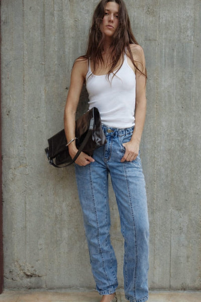 Woman wearing a white tank top and blue jeans, holding a black bag against a concrete wall.
