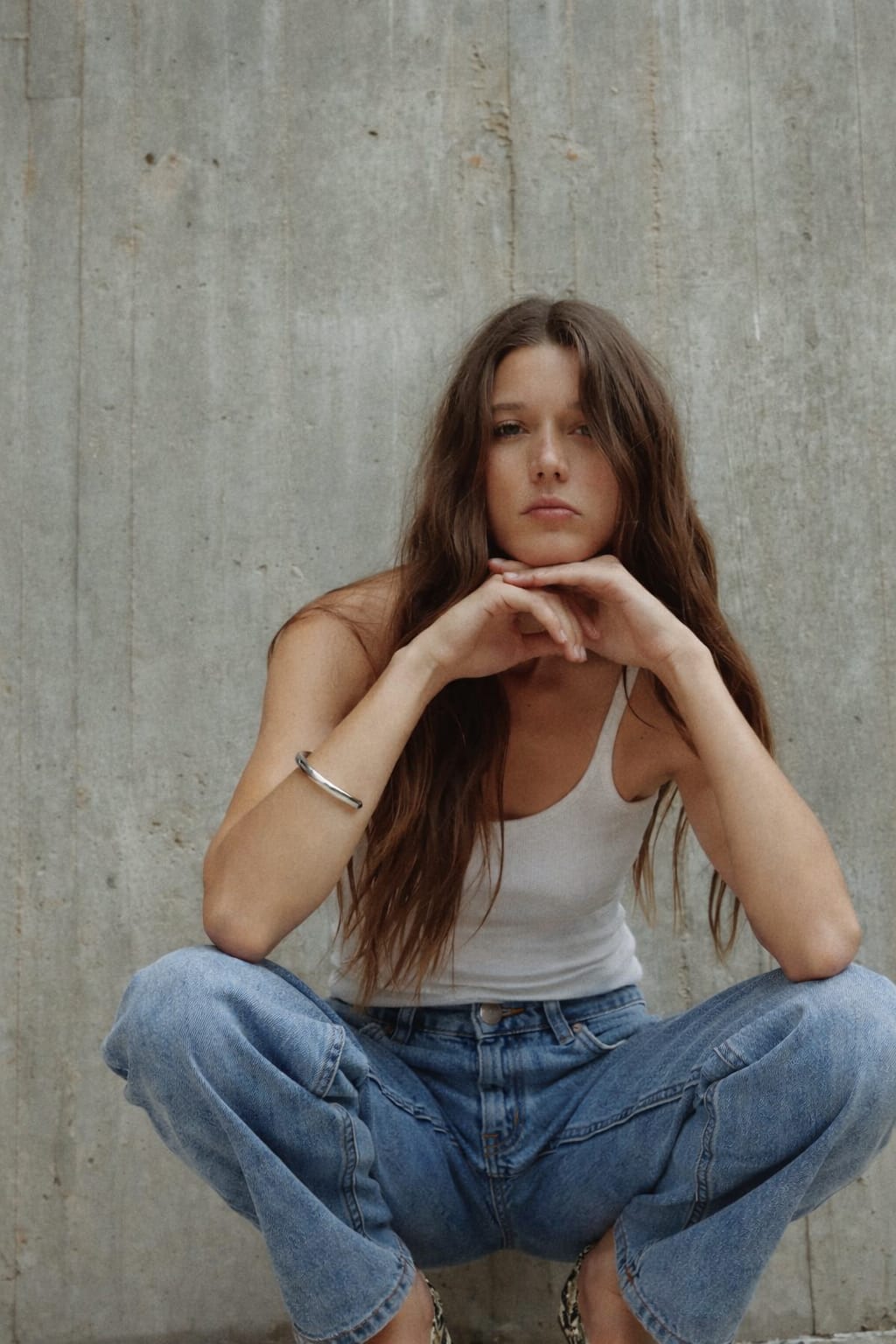 Woman sitting against a concrete wall wearing a white tank top and blue jeans.