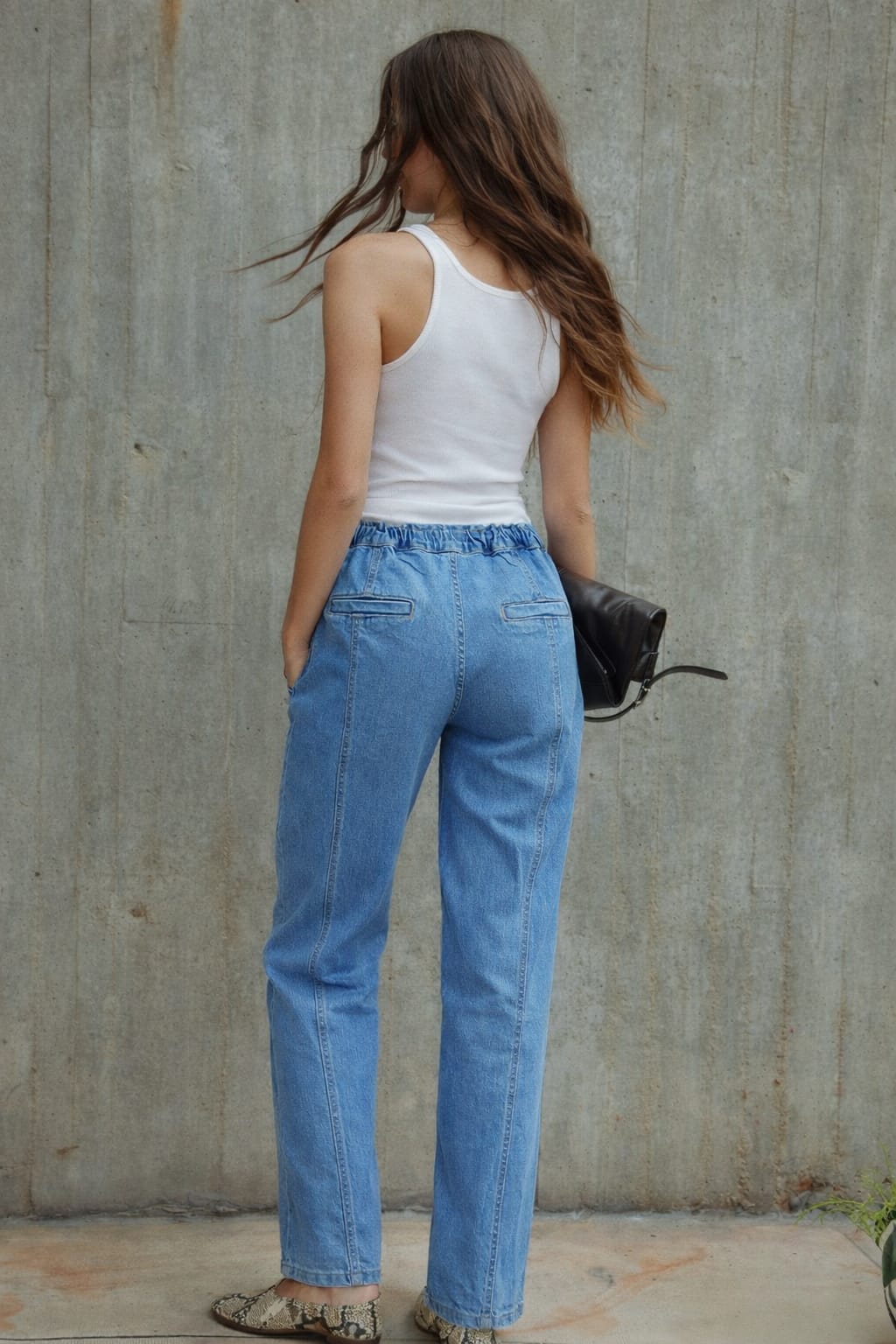 Woman wearing a white tank top and blue jeans standing against a concrete wall.