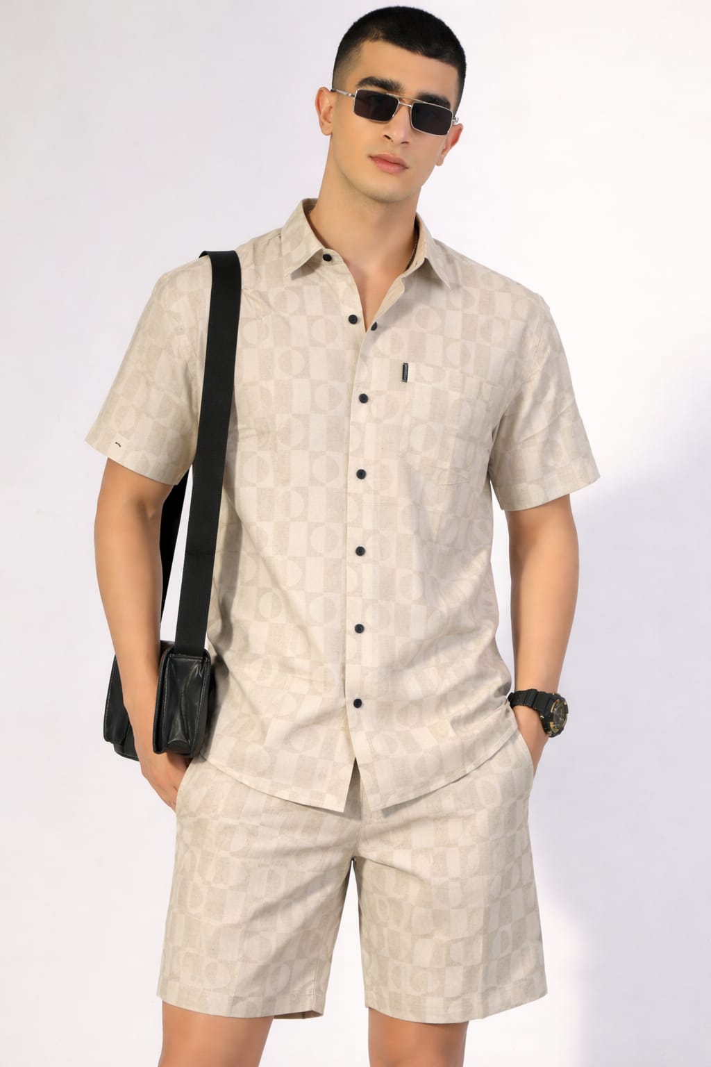 Man wearing a beige patterned shirt and shorts with sunglasses and a black bag on a white background