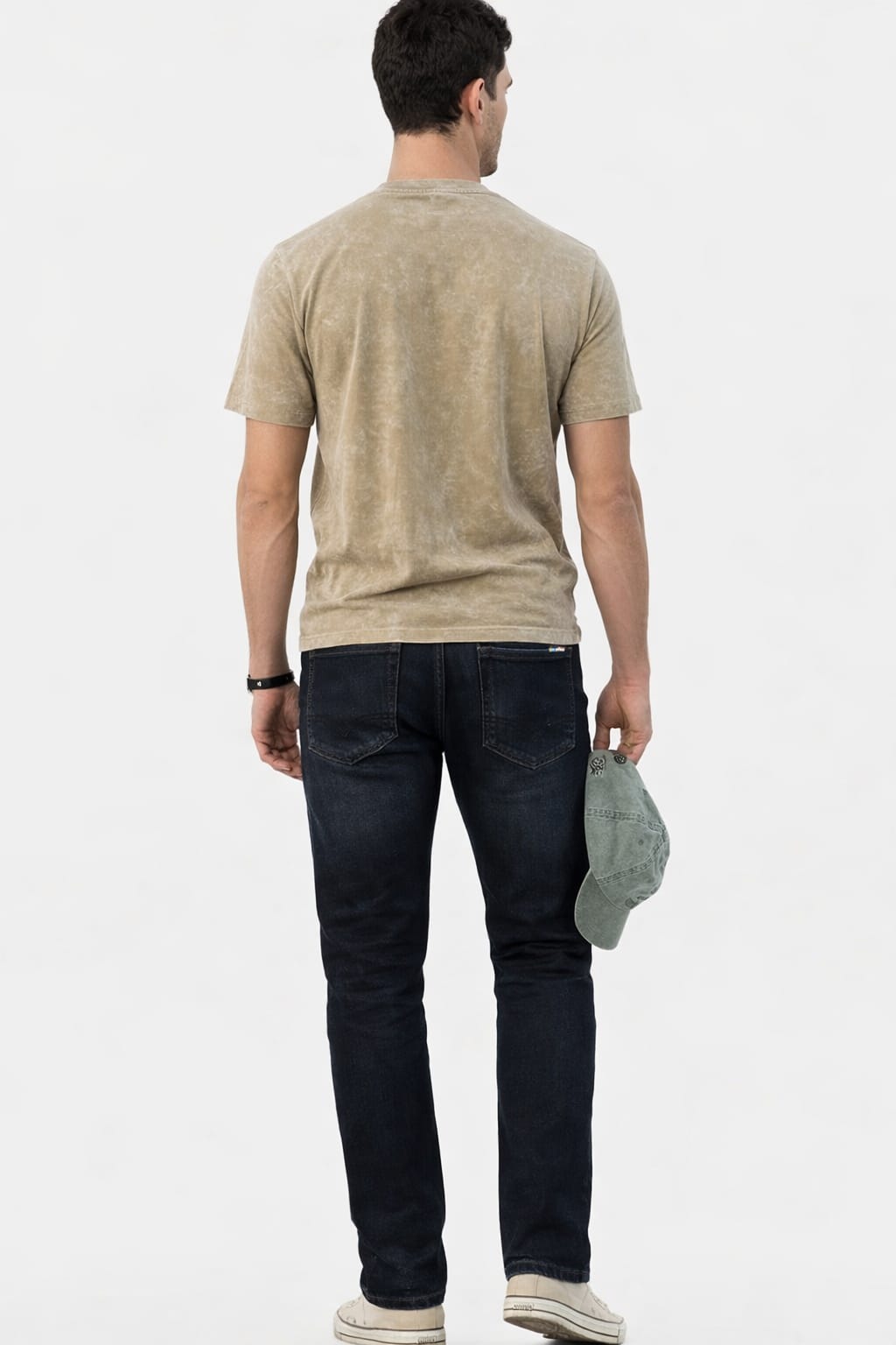 Man wearing a beige t-shirt and dark jeans holding a cap on a white background