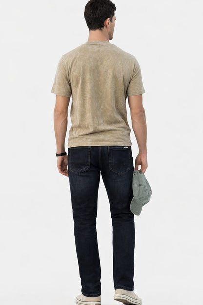 Man wearing a beige t-shirt and dark jeans holding a cap on a white background