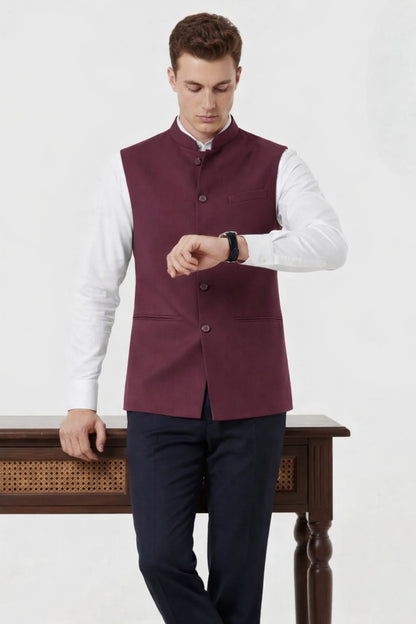 Man wearing a maroon vest over a white shirt with dark pants, standing against a white background.