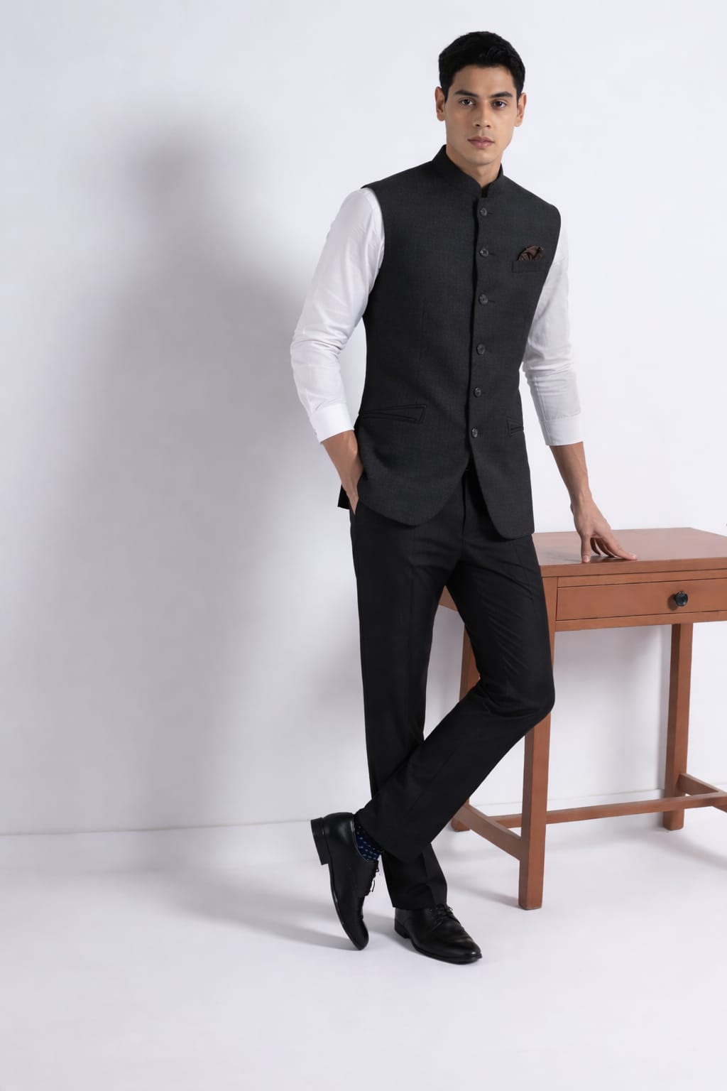 Man wearing a black vest over a white shirt with black pants, standing against a white background.