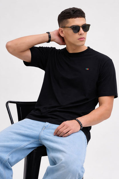 Man wearing a black t-shirt and light blue jeans sitting on a chair with a white background
