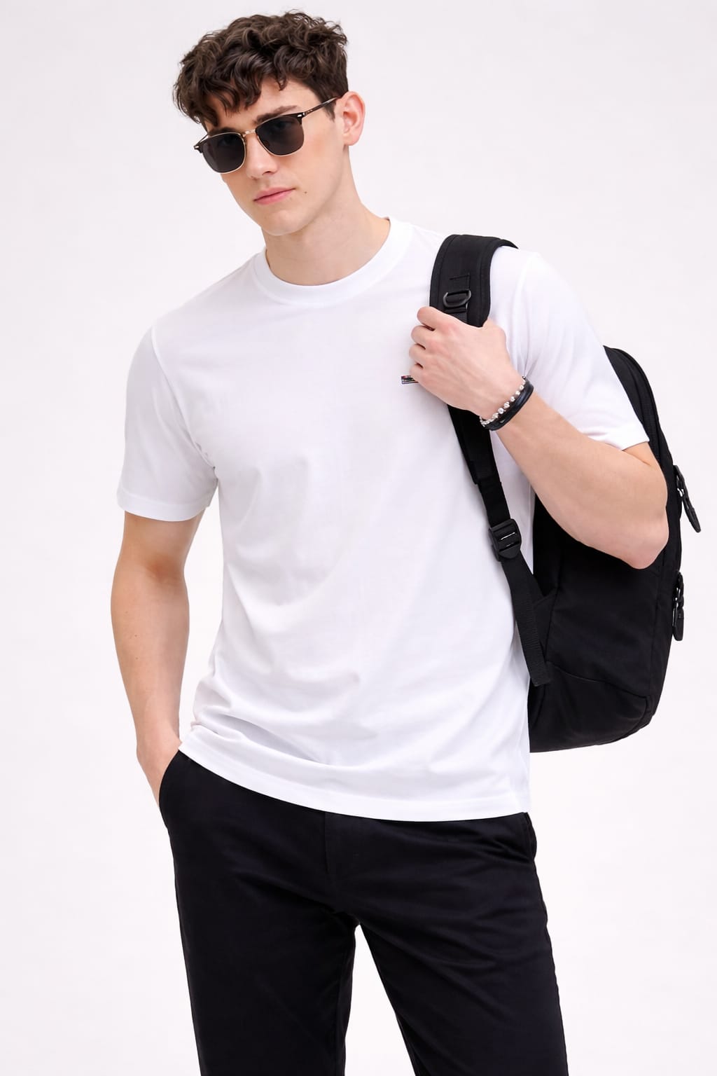 Man wearing a white t-shirt and black pants, holding a black backpack against a white background