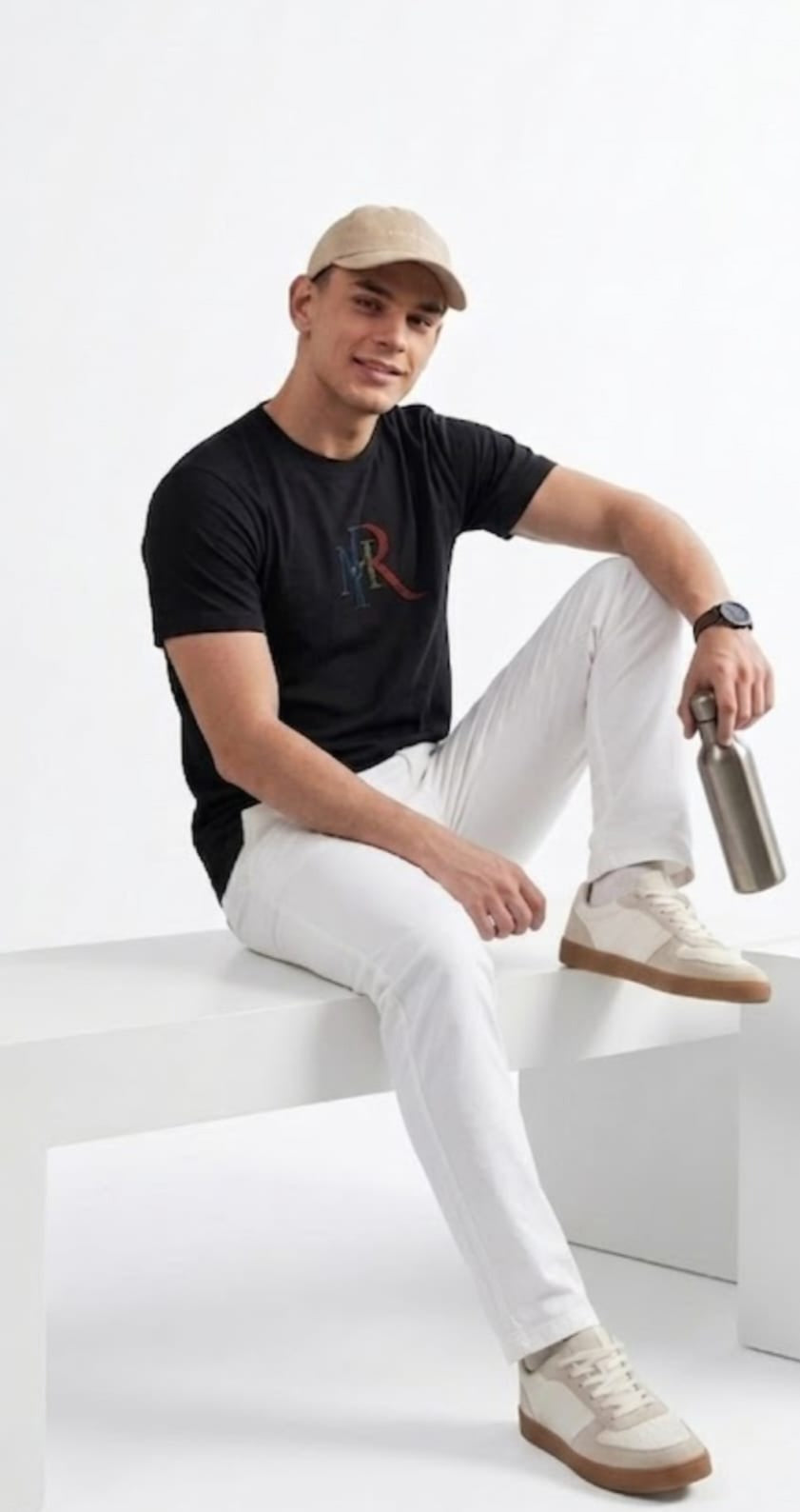 Man sitting on a white cube wearing a black t-shirt with a logo, white pants, and beige cap, holding a silver water bottle.