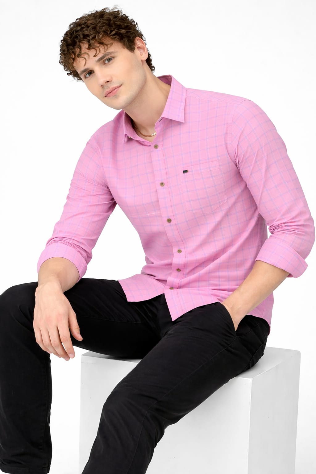 Man wearing a pink shirt and black pants sitting on a white stool against a white background