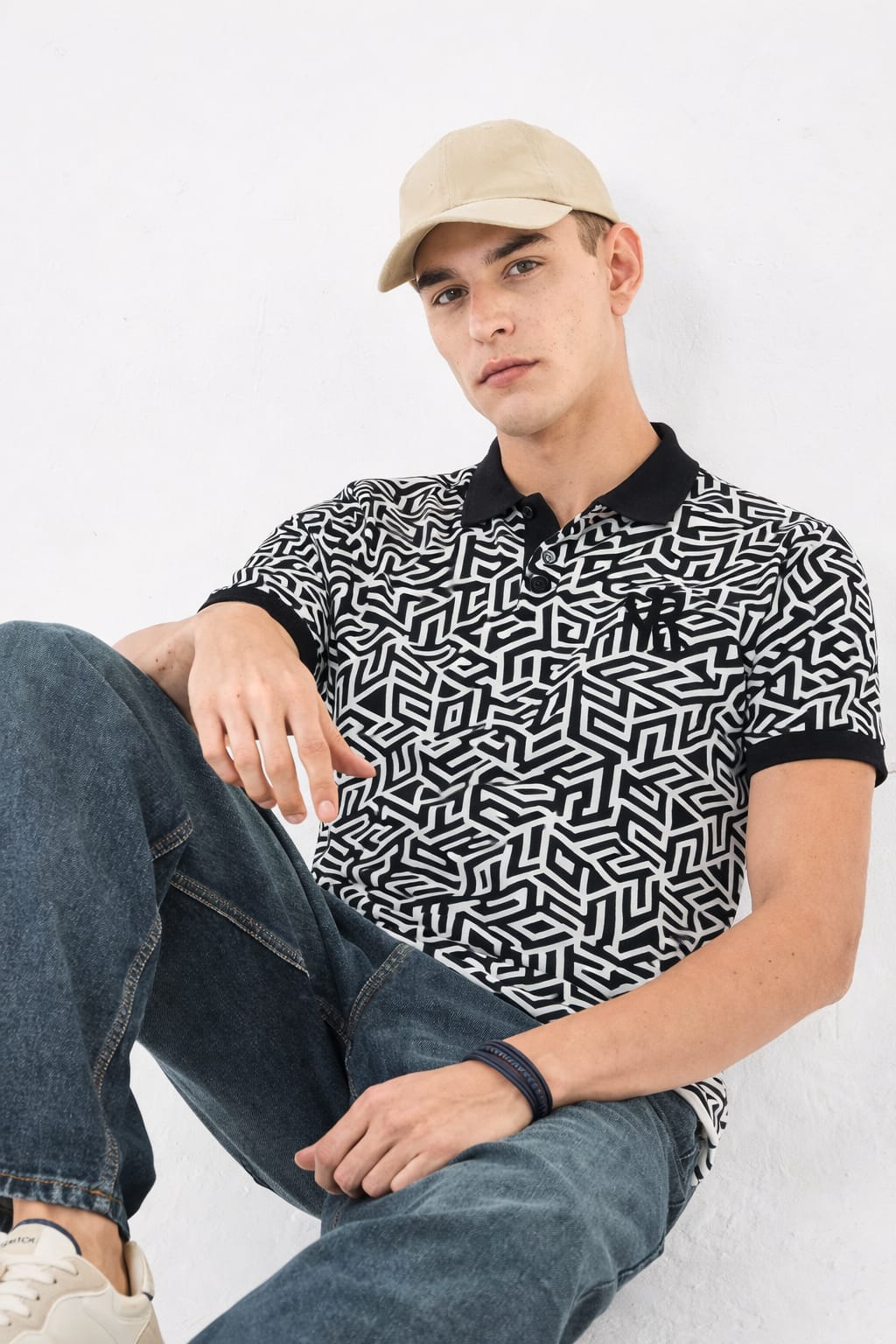 Man wearing a black and white geometric patterned polo shirt with a beige cap and jeans, sitting on a white background.