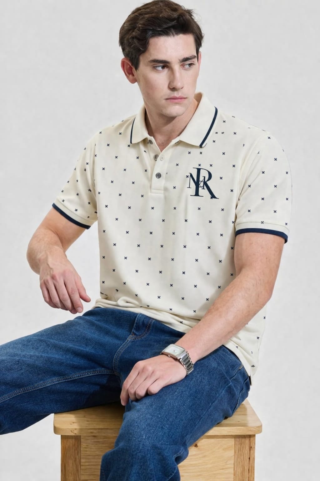 Man wearing a patterned polo shirt with a logo, sitting on a wooden stool against a plain background