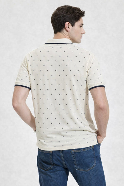 Man wearing a cream-colored polo shirt with black patterns and blue jeans on a white background