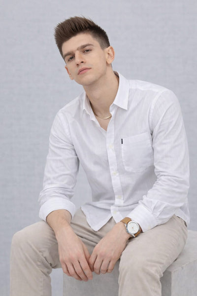 Man wearing a patterned polo shirt with a logo, sitting on a wooden stool against a plain background