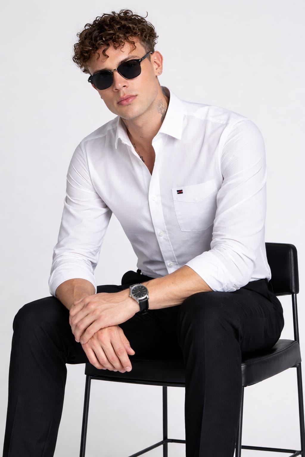 Man wearing a white shirt and black pants sitting on a chair against a plain background