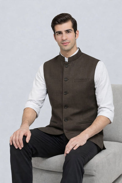 Man wearing a brown vest over a white shirt and black pants, sitting on a gray couch against a light gray background.