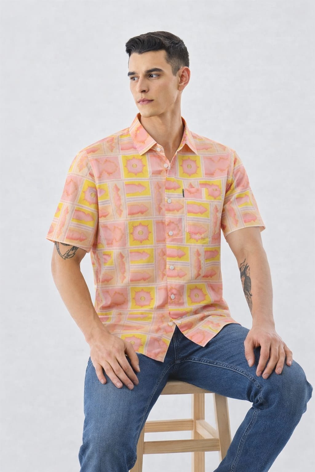 Man wearing a patterned pink shirt and blue jeans sitting on a stool against a white background
