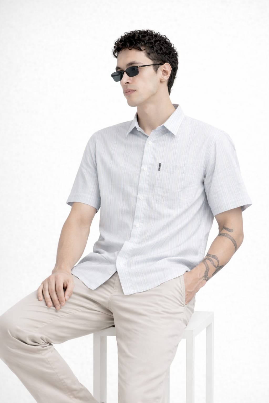 Man wearing a light gray short-sleeve shirt and beige pants sitting on a white stool against a white background.